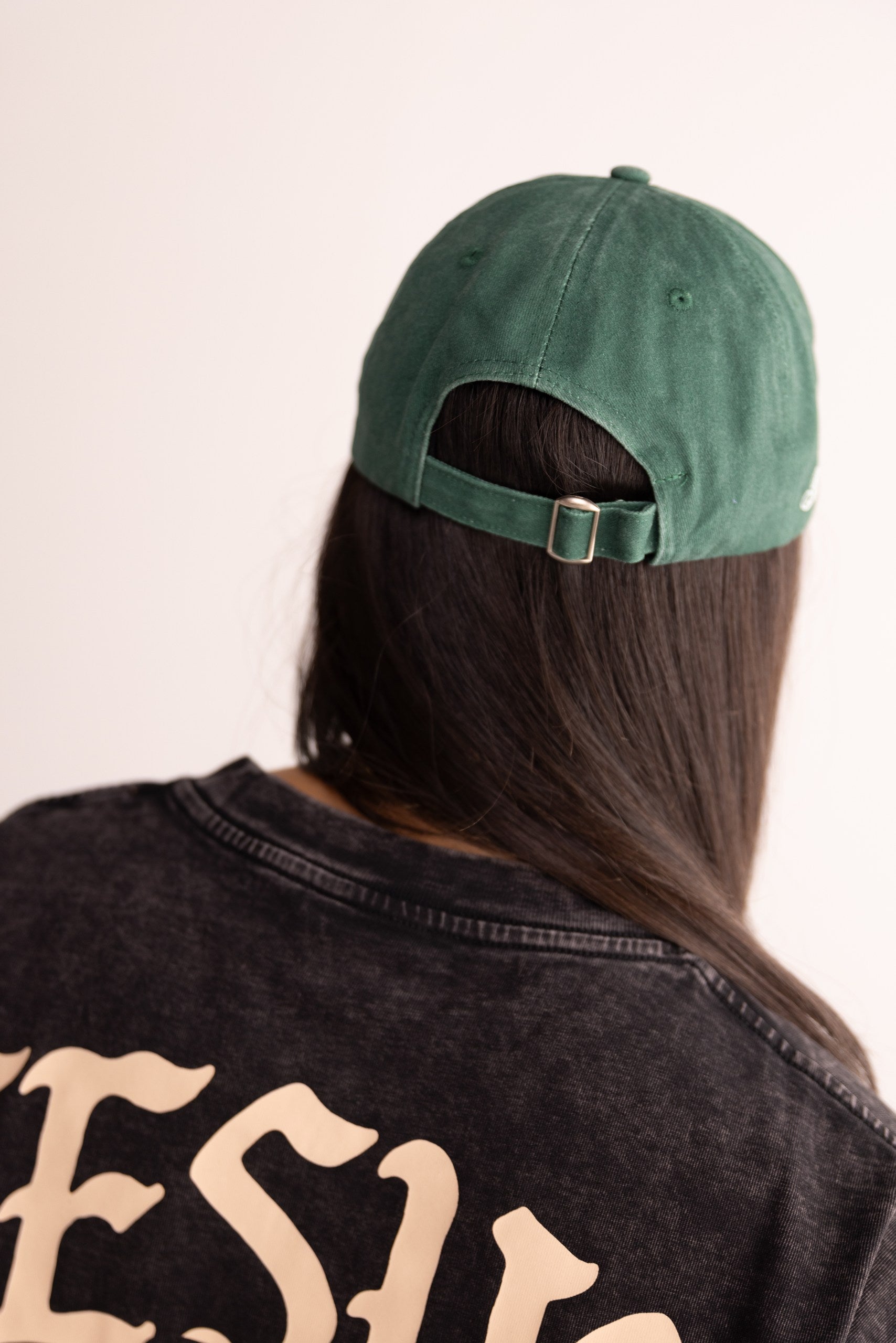 Person wearing a green cap and black jacket with 'Jesus' on the back, against a plain background