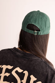 Person wearing a green cap and black jacket with 'Jesus' on the back, against a plain background