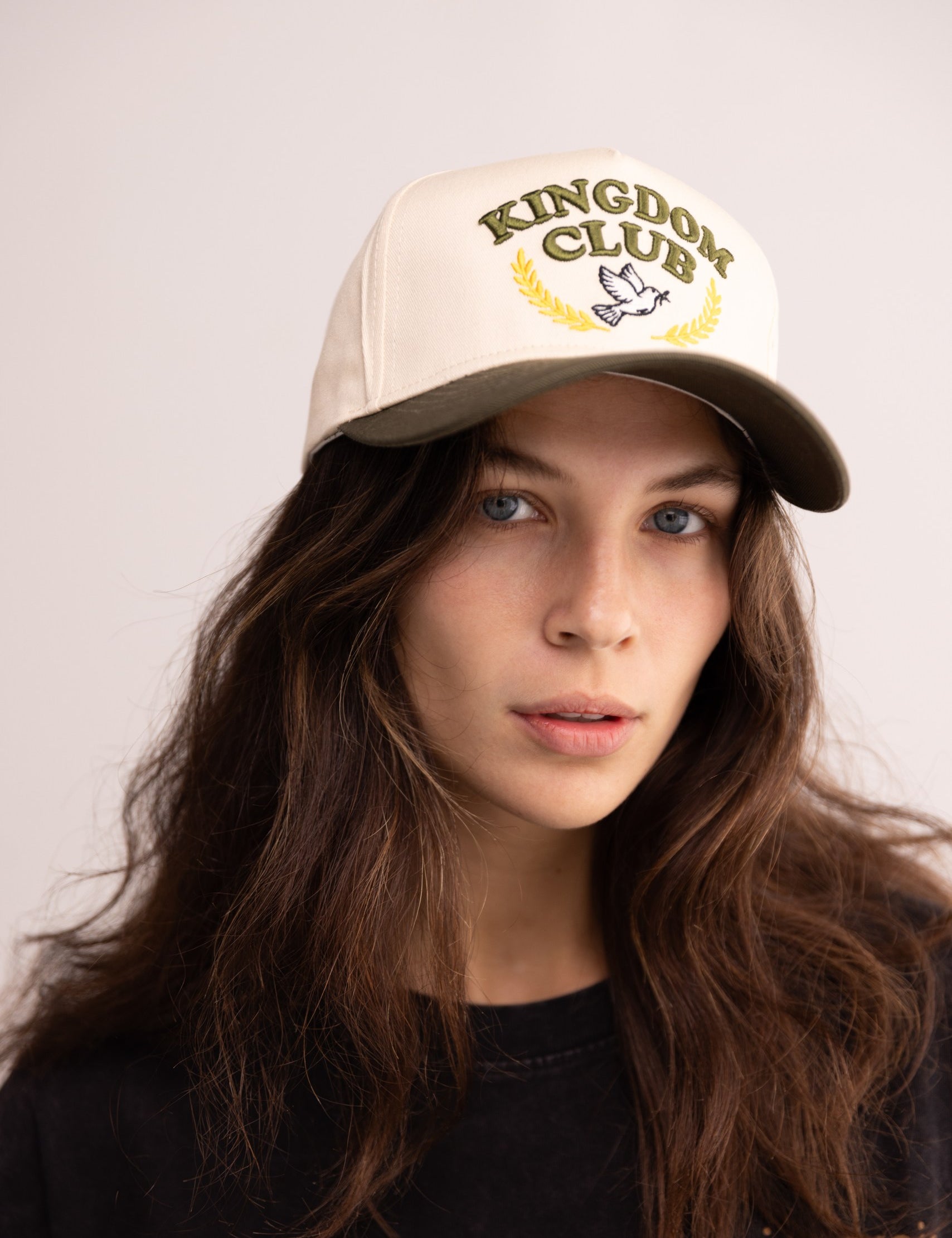 Woman wearing a beige cap with 'Kingdom Club' logo on a plain background