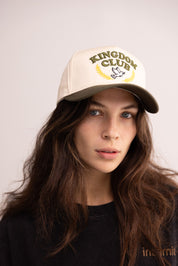 Woman wearing a beige cap with 'Kingdom Club' logo on a plain background