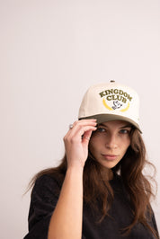 Person wearing a beige cap with 'Kingdom Club' logo on a plain background