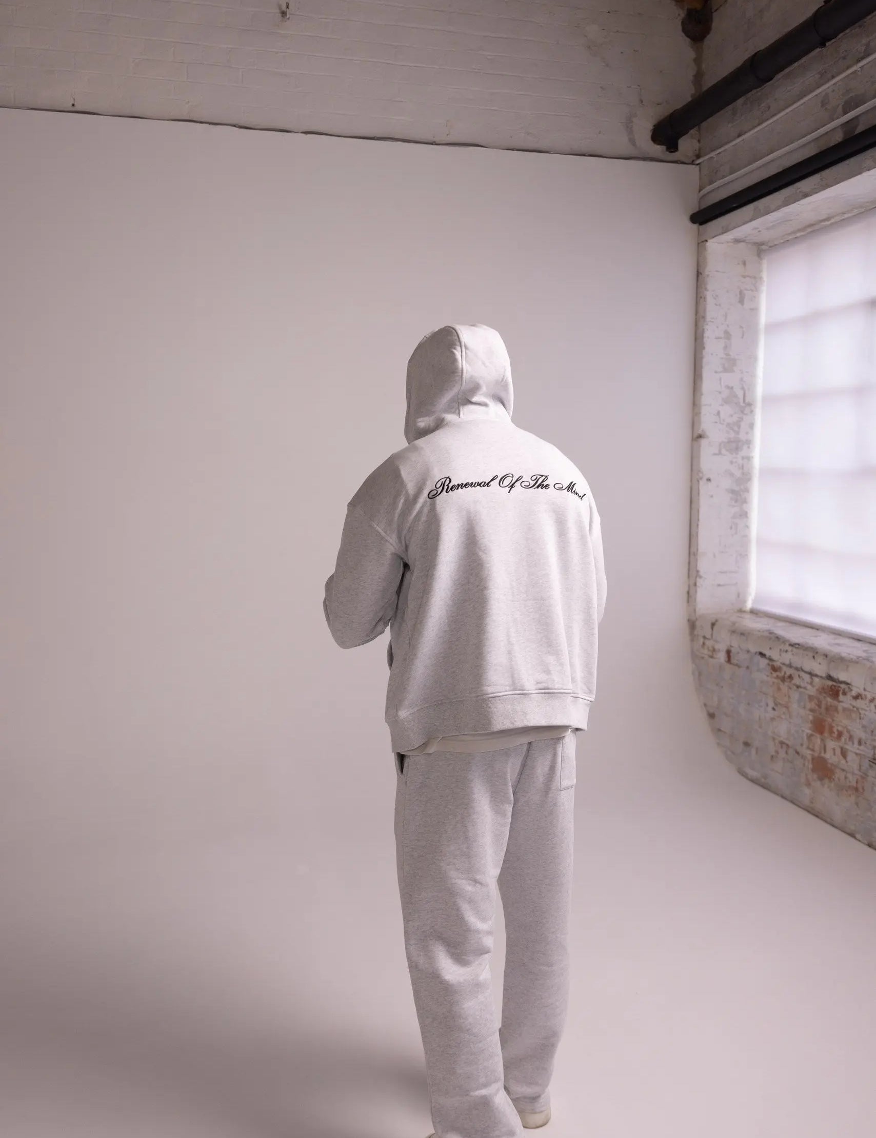 Person wearing a grey hoodie and pants in a minimalistic room with white walls and a brick wall.