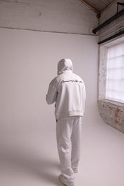 Person wearing a grey hoodie and pants in a minimalistic room with white walls and a brick wall.