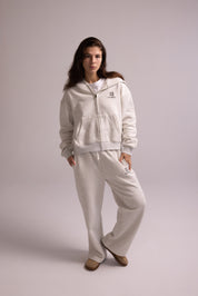 Woman wearing a light gray tracksuit with a logo on a plain background